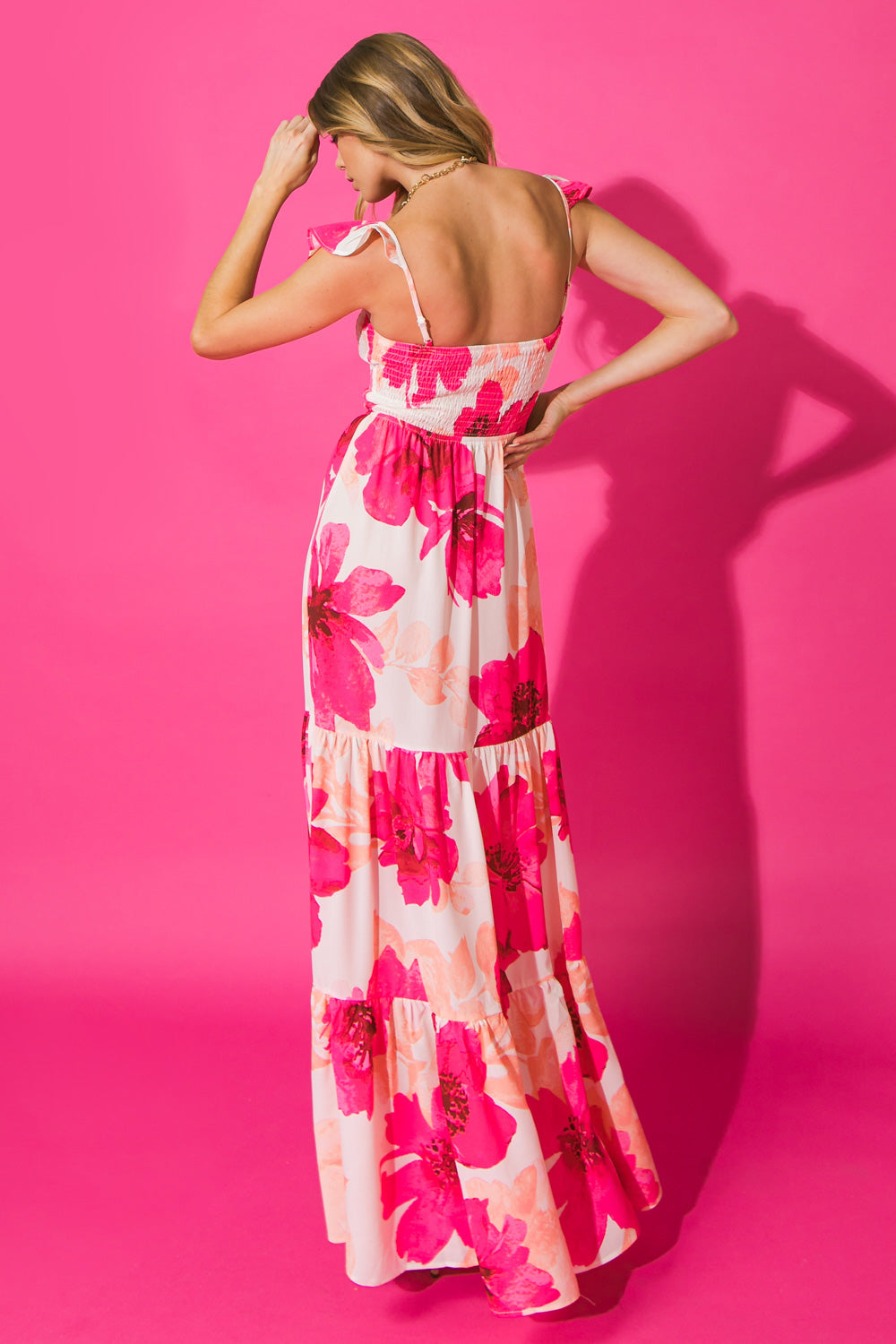 A woman stands wearing a long, floral-patterned dress with pink flowers. She touches her hair, facing away from the camera, against a bright pink background, creating a vibrant, stylish look.