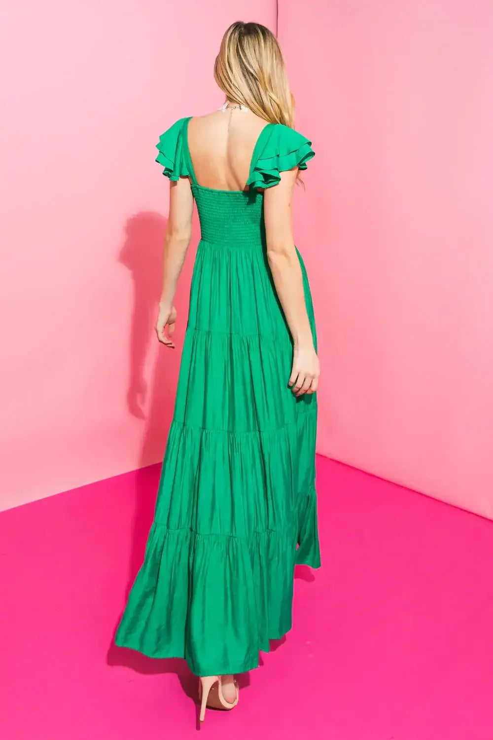 A woman in a green, ruffled maxi dress stands with her back to the viewer against a pink backdrop, showcasing the dress's tiered design and flowing fabric.