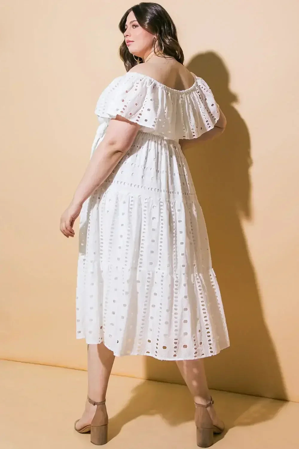A woman stands in a white, off-shoulder dress with eyelet detailing, facing slightly away. She is posed against a soft, beige background, showcasing her elegant silhouette.