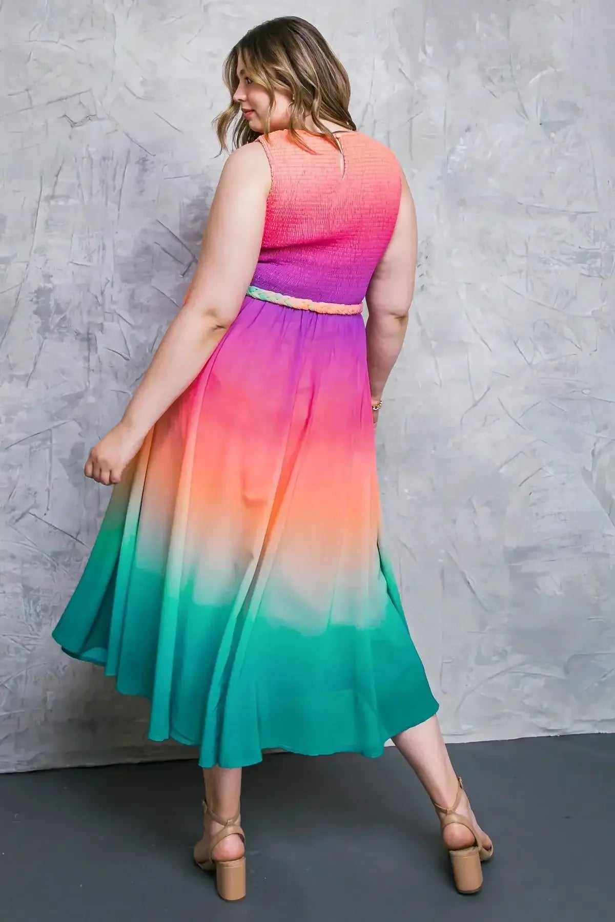 A woman wearing a colorful, gradient dress is looking over her shoulder. The dress transitions from pink to orange to green. She stands against a textured gray wall.