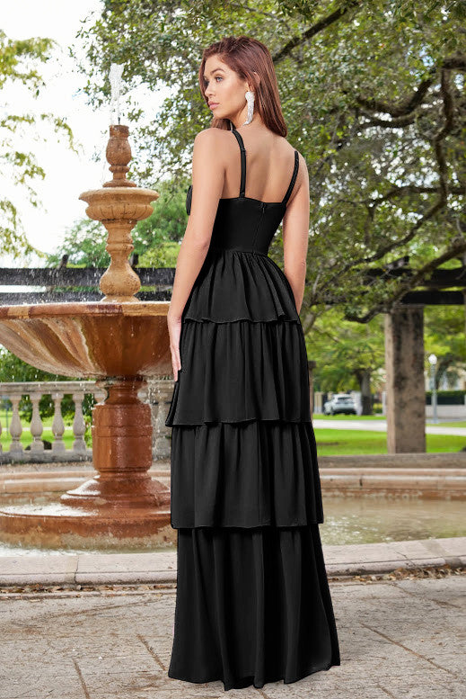 A woman in a tiered black dress stands in front of a stone fountain, surrounded by greenery and paved pathways in a park-like setting, appearing elegant and poised.