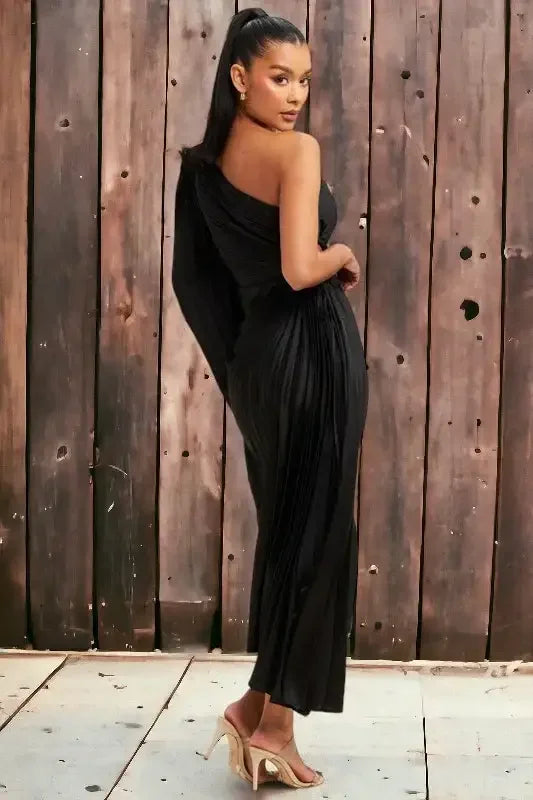 A woman is posing gracefully in a black, one-shoulder pleated dress, with her hair pulled back. She stands against a rustic wooden backdrop, accentuating a chic, elegant atmosphere.