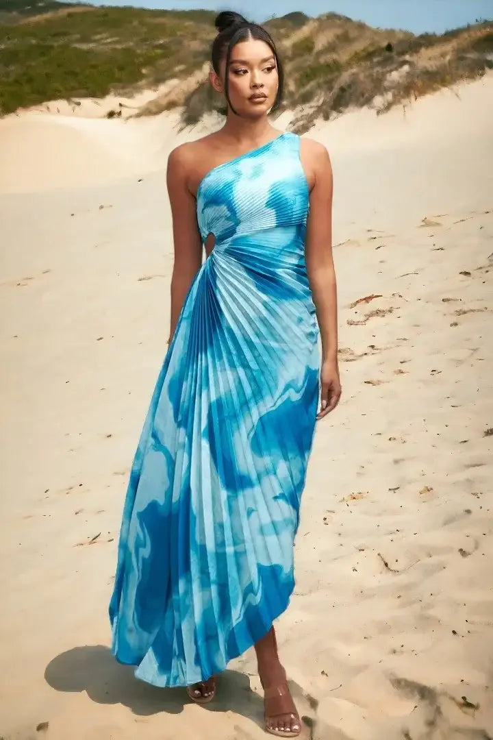 A model stands on sandy terrain, wearing a flowing blue dress with a one-shoulder design and pleated texture, set against a backdrop of gentle hills and greenery.