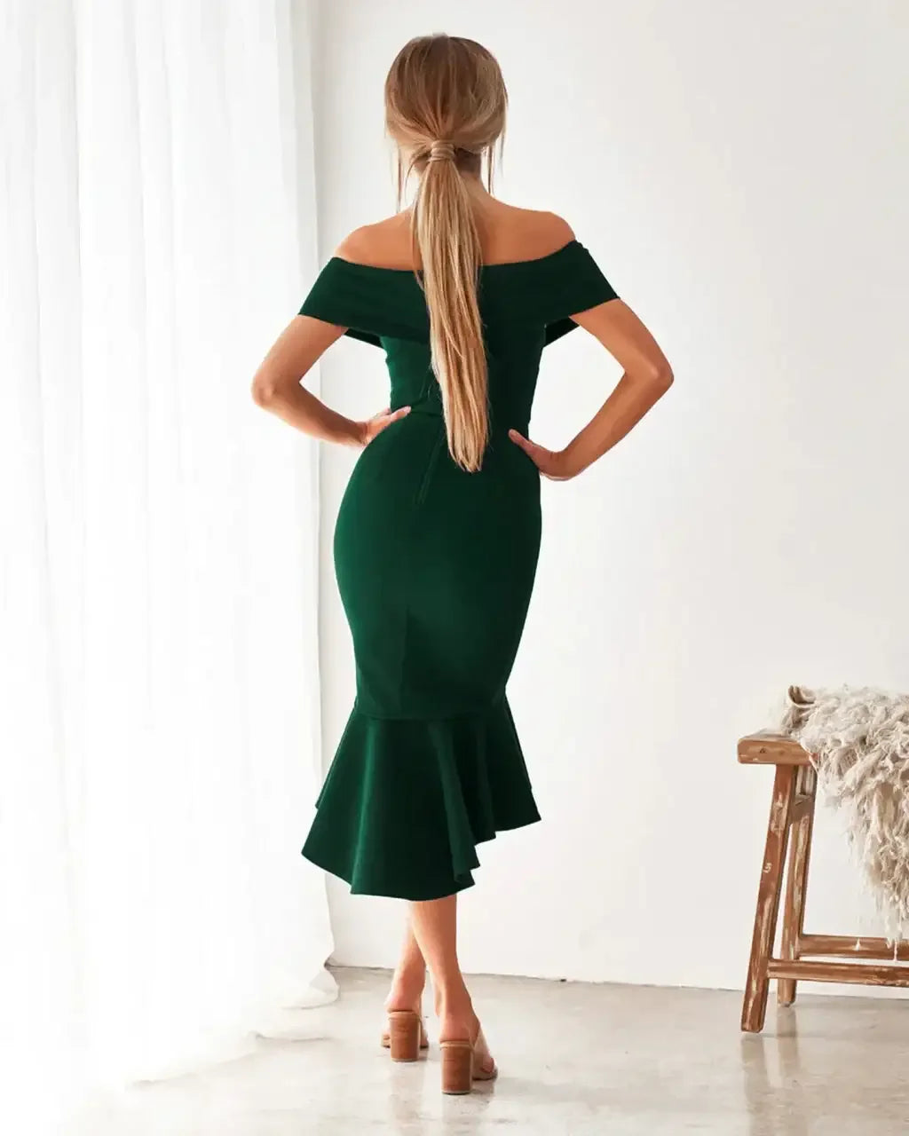 A woman stands with her back to the viewer, wearing a green off-shoulder dress with a flared hem. Natural light filters through sheer curtains in a minimalistic indoor setting.