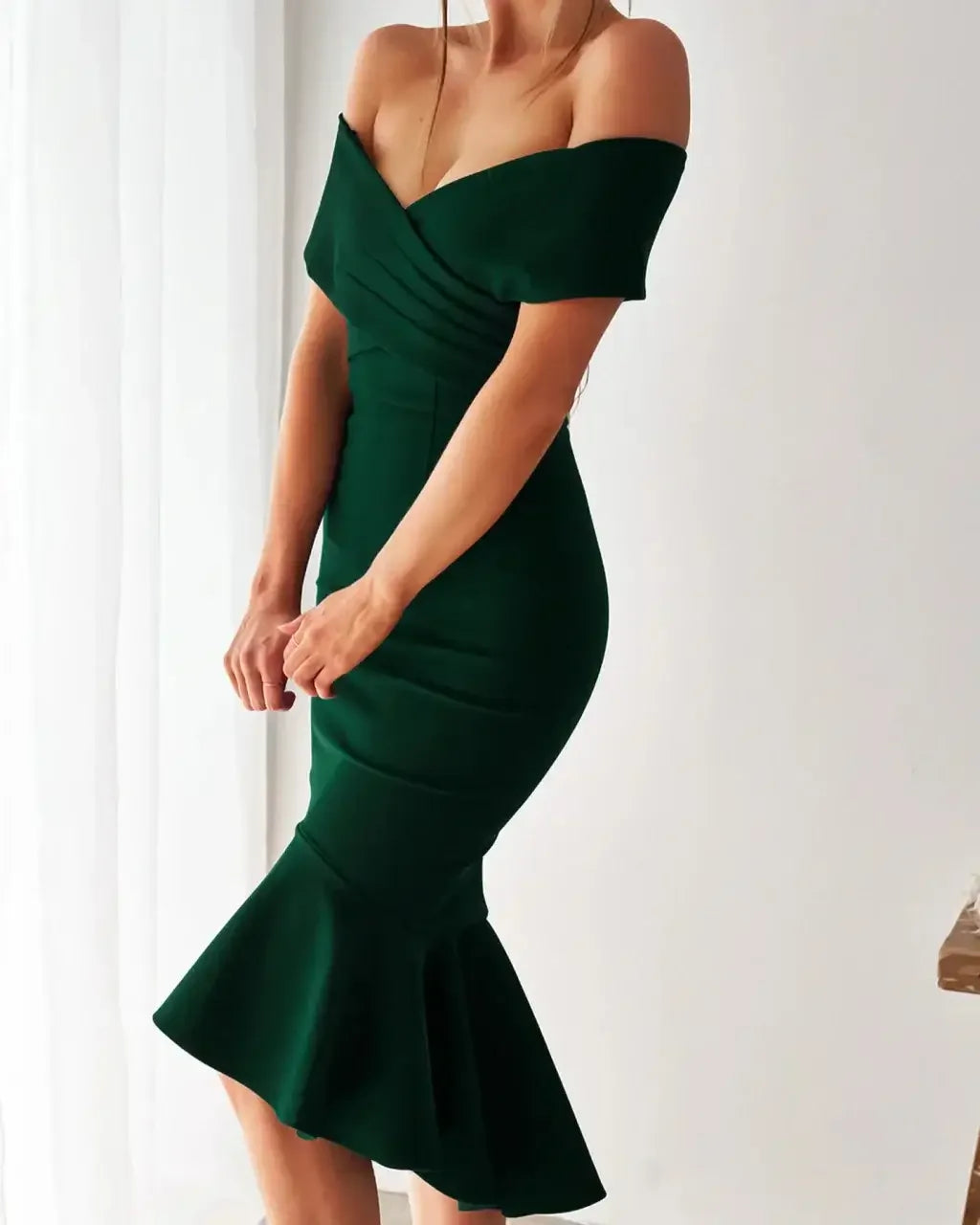 A woman wears a green off-shoulder dress with a fitted bodice and a flared hem. She is posing with her hands near her waist, in a softly lit, neutral-toned interior.