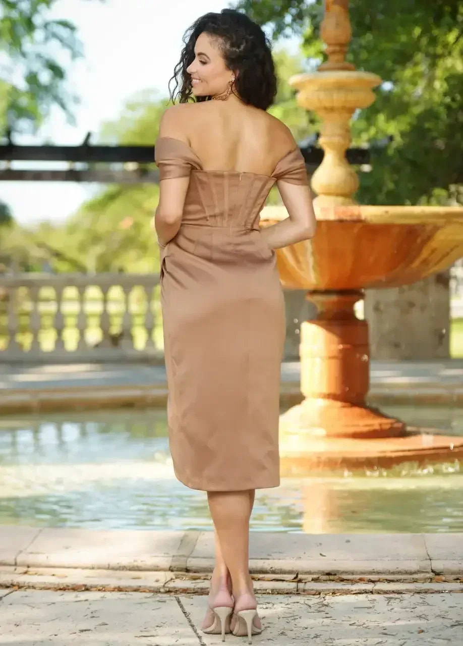 A woman stands gracefully in a fitted, off-shoulder, beige dress. She looks back, smiling, in a park setting with a fountain and lush greenery in the background.