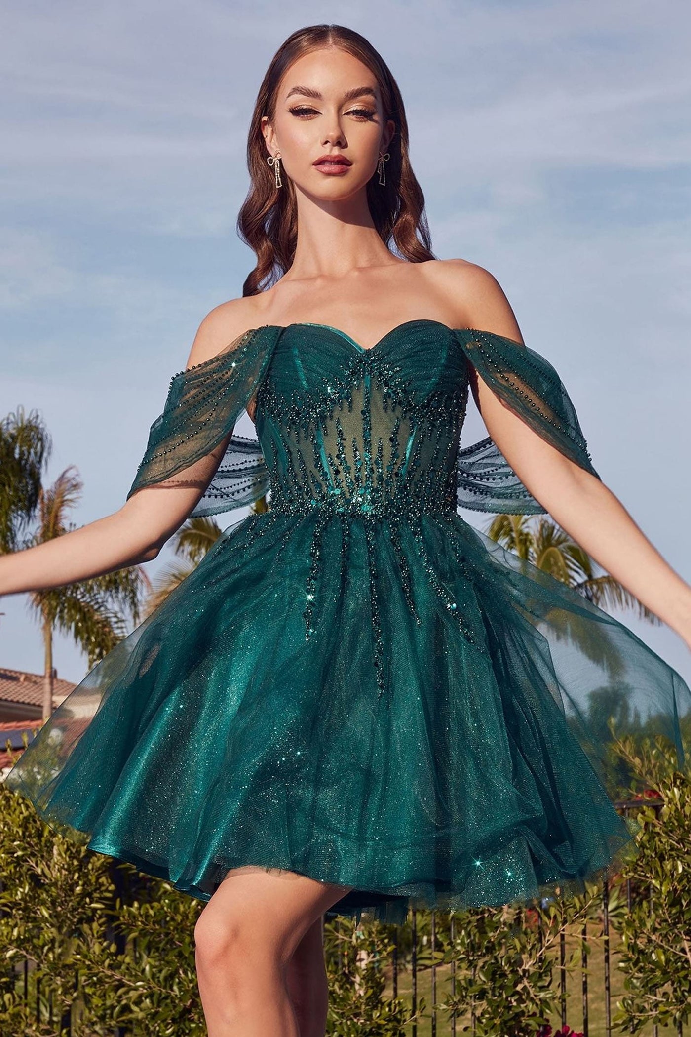 A woman wearing a sparkling off-the-shoulder mini dress in deep teal green, standing outdoors with her arms outstretched against a sky and palm trees background.