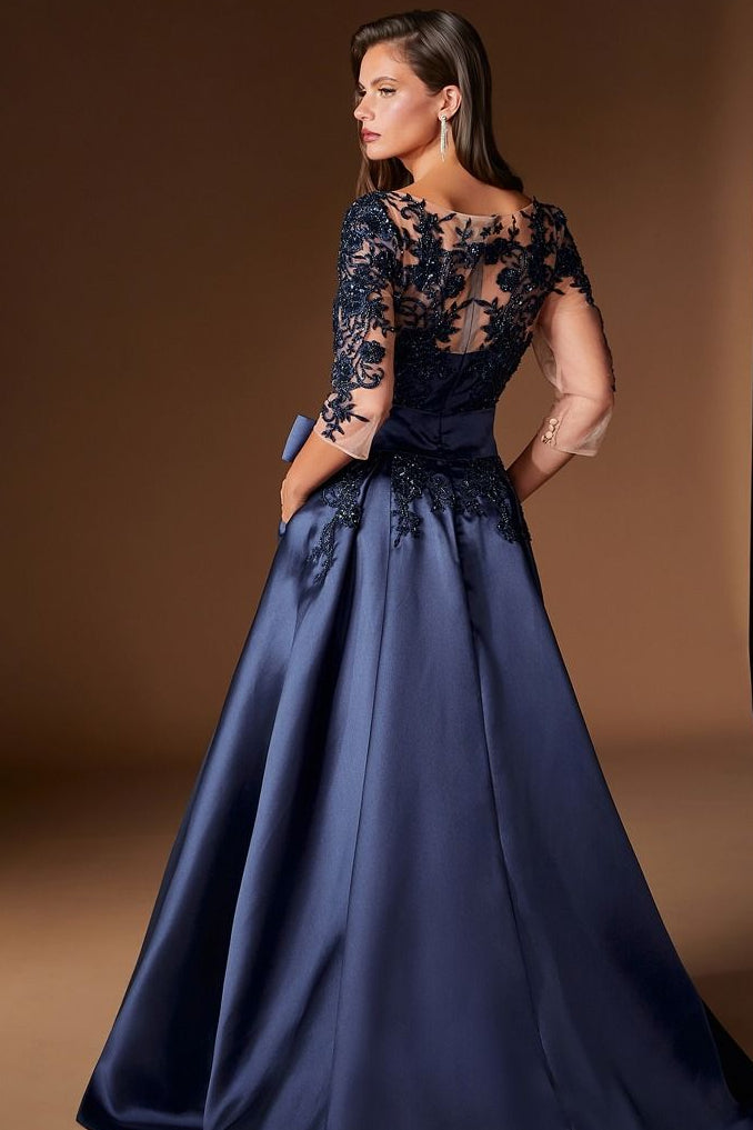 A woman modeling a floor-length navy blue gown with a sheer lace bodice, short sleeves, and a full satin skirt, photographed in a studio with a warm brown backdrop.