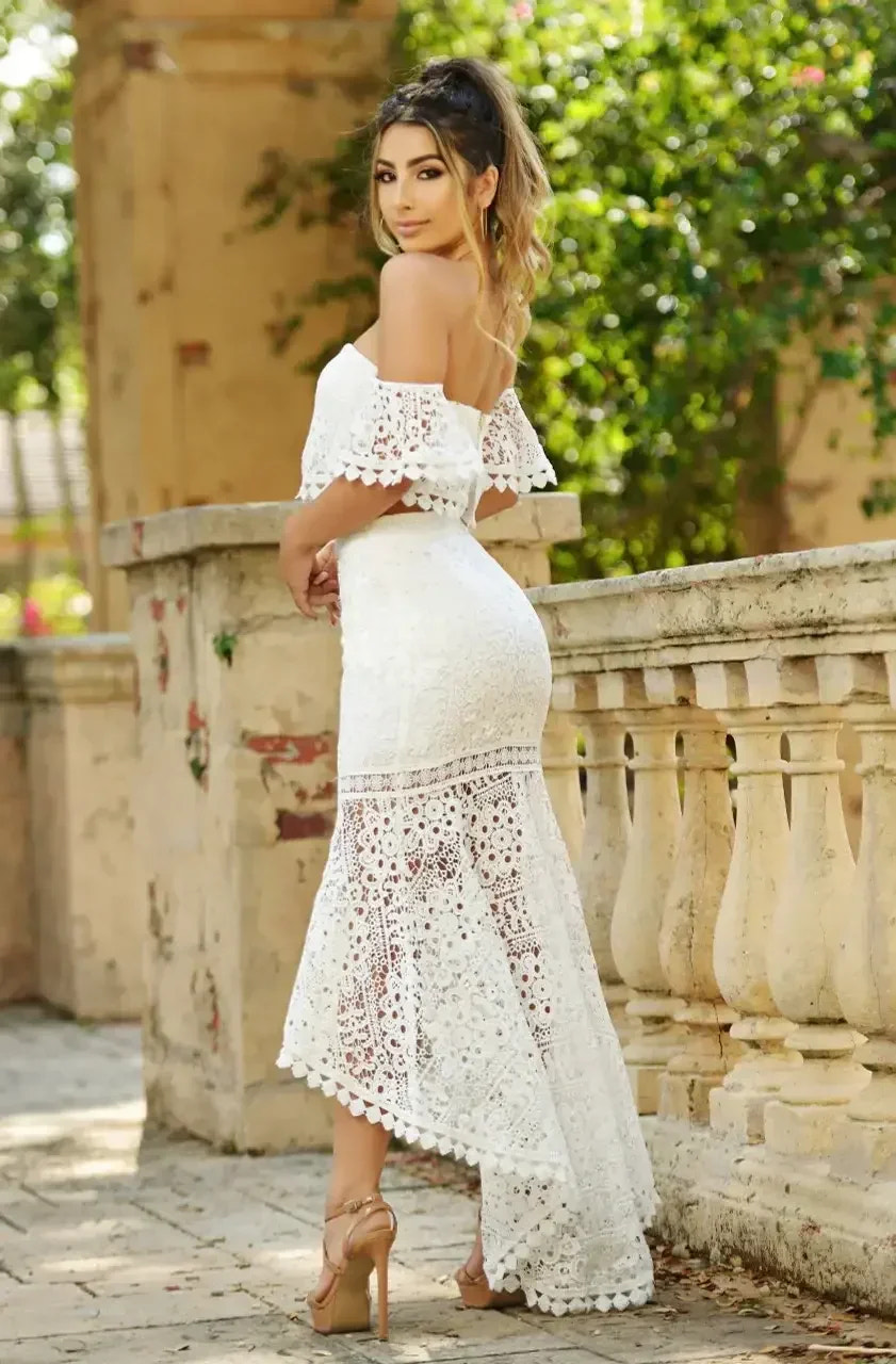 A woman in a white lace dress poses confidently, her hair in loose curls. She stands near a stone railing surrounded by greenery, illuminated by soft sunlight.