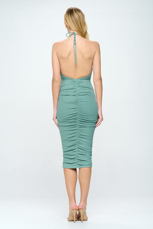 A fitted, green midi dress with ruching along the back is worn by a woman, showcasing her bare shoulders. The neutral background emphasizes the dress's color and design.