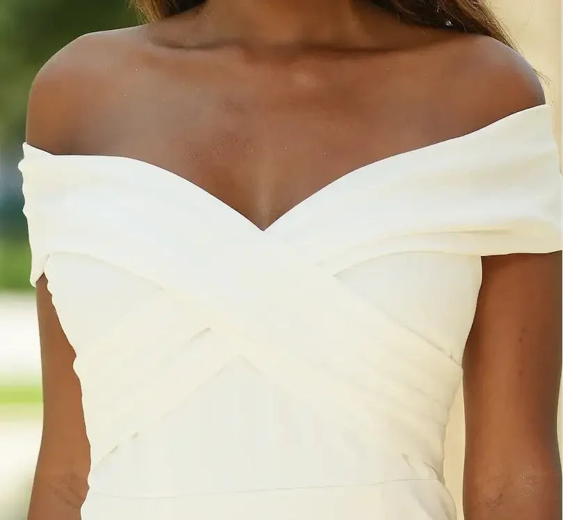 A white off-shoulder dress elegantly features crossed fabric at the chest. The focus is on the dress's neckline against a blurred outdoor background, suggesting a sunny, relaxed setting.
