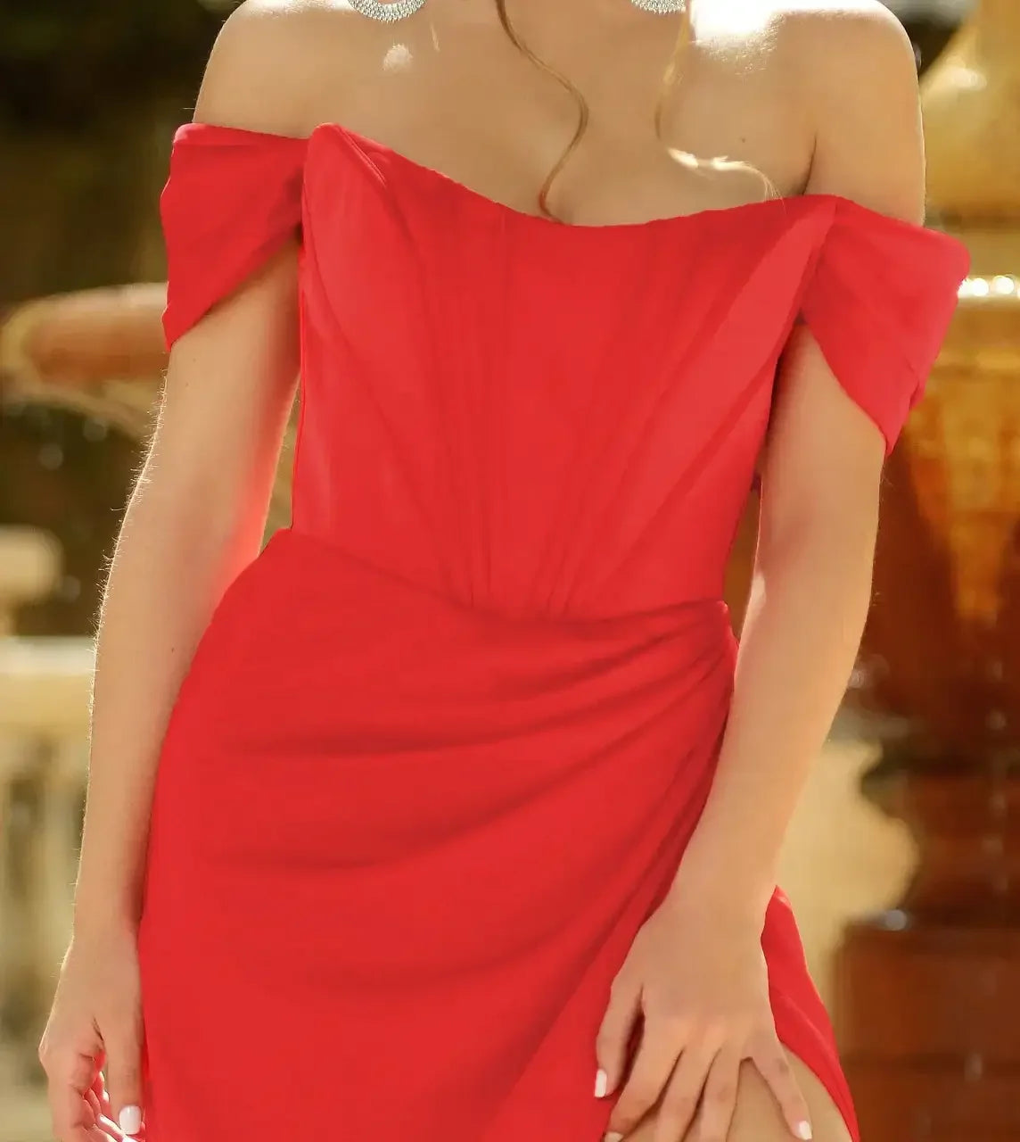 A woman is wearing a bright red, off-shoulder dress with pleats, holding her hand near the hem. The setting features a blurred background of a decorative fountain.