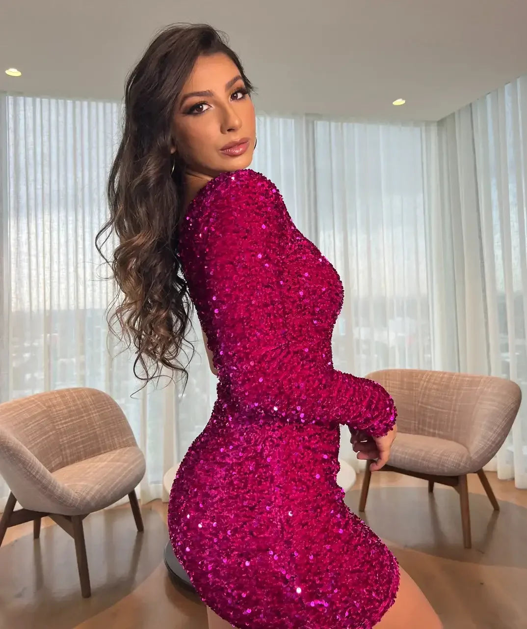 A woman poses in a sparkling magenta dress, showcasing her figure. She is in a well-lit indoor space with large windows and modern chairs, creating an elegant atmosphere.