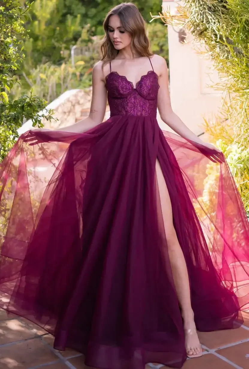 A woman in a flowing burgundy dress twirls, showcasing a high slit and delicate lace details, amidst a lush garden with vibrant greenery in the background.