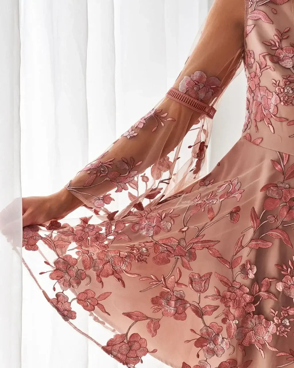 A hand delicately holds the skirt of a flowy, embroidered dress, showcasing floral patterns against a soft, sheer fabric. The backdrop features softly illuminated, sheer curtains.