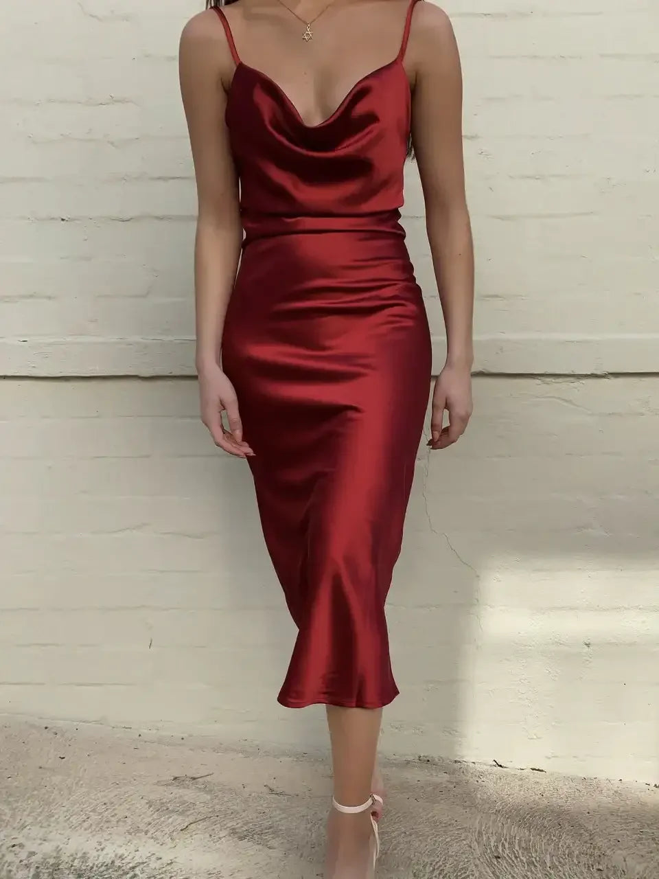 A person stands wearing a sleek, red satin dress with thin straps, showcasing a fitted silhouette. The background features a light-colored, textured wall, adding elegance to the scene.