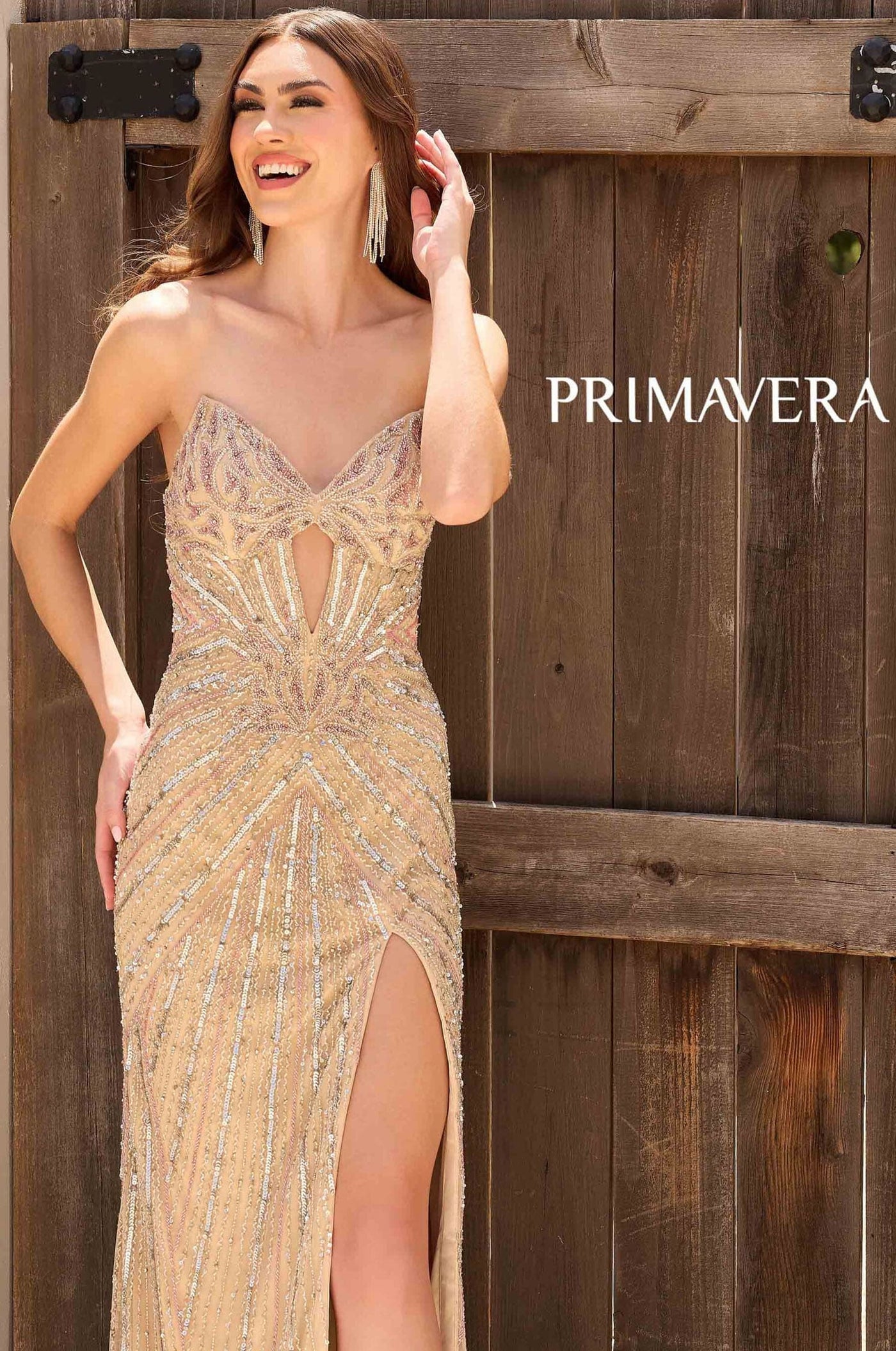 Woman in a beaded evening gown standing against a wooden background with 'PRIMAVERA' branding.