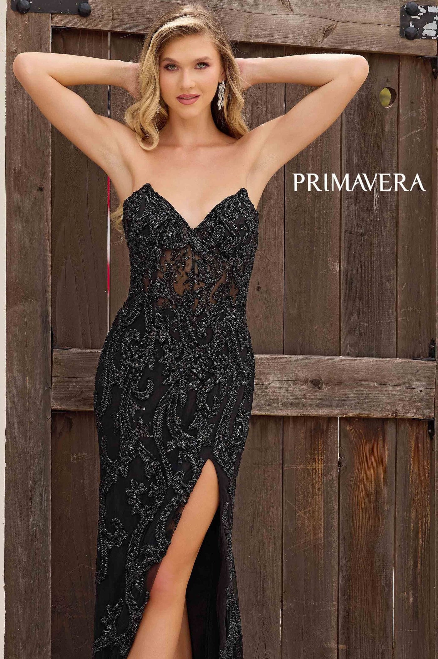 Woman wearing a black lace dress with a high slit, standing against a wooden background with 'PRIMAVERA' branding.