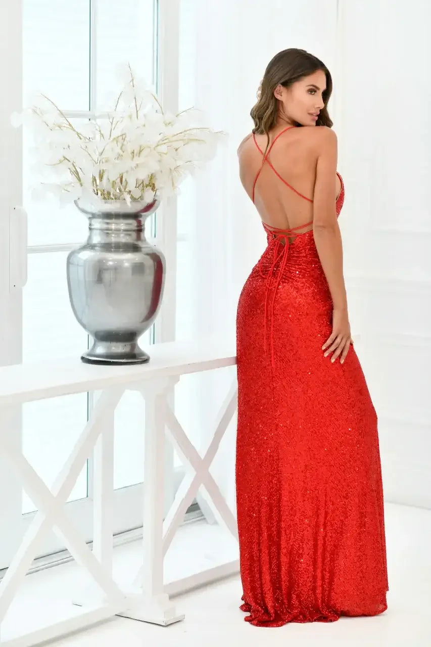 A woman in a stunning red sequined gown stands gracefully, displaying an elegant back design. She poses beside a silver vase filled with white flowers in a bright, minimalistic room.