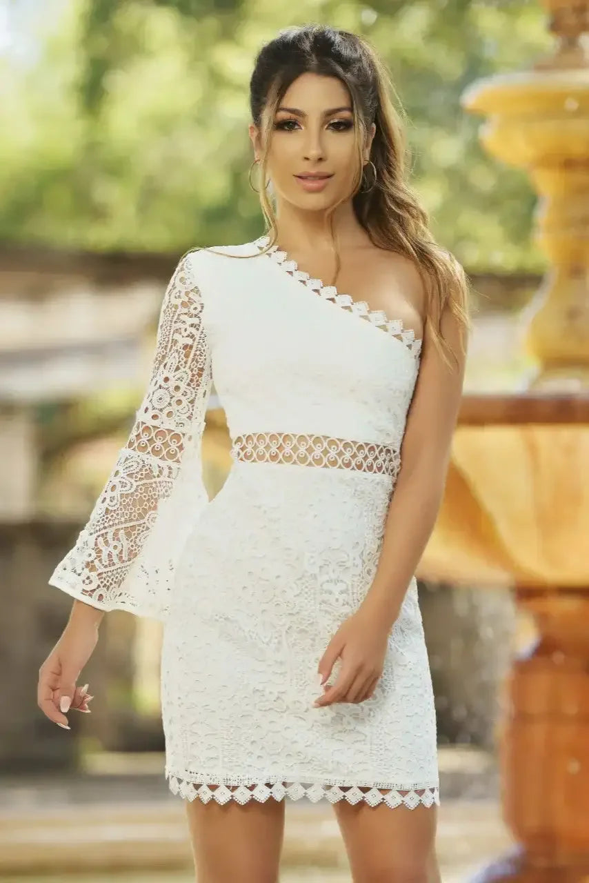 A woman wears a stylish white dress with lace details, standing confidently by a fountain in a lush green outdoor setting. Sunlight filters through the trees, enhancing the scene.