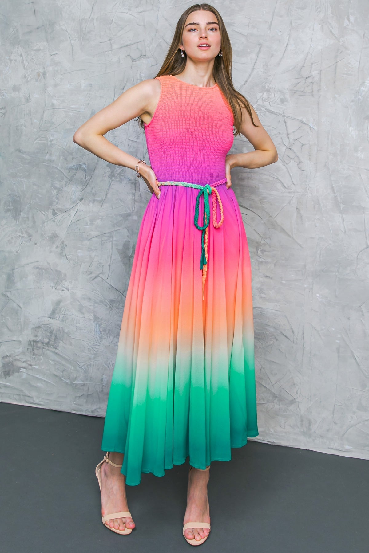 A woman poses confidently in a colorful, gradient dress transitioning from pink to green, highlighting her long hair against a textured gray wall, with a subtle focus on her stylish sandals.