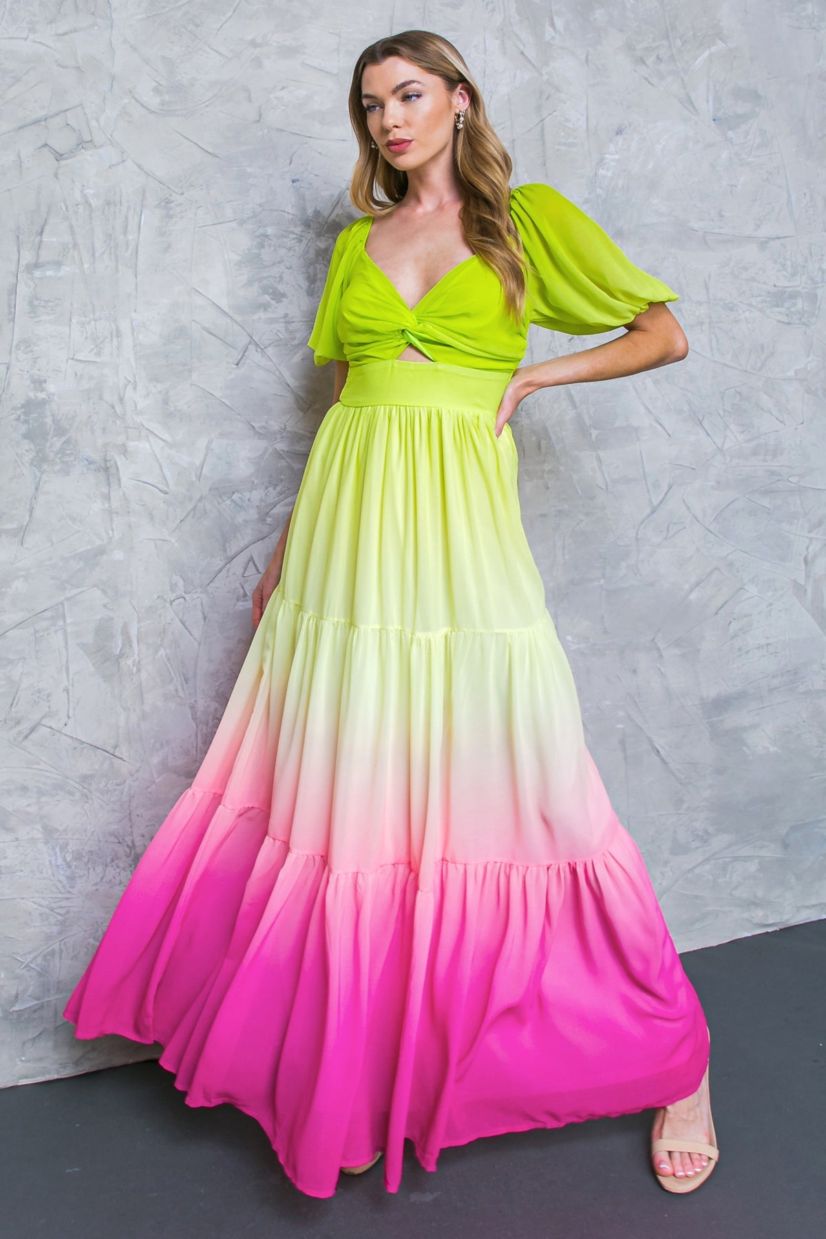A woman in a vibrant, tiered maxi dress transitions from bright green to pink. She poses confidently against a textured gray wall, showcasing the dress’s flow and color gradient.