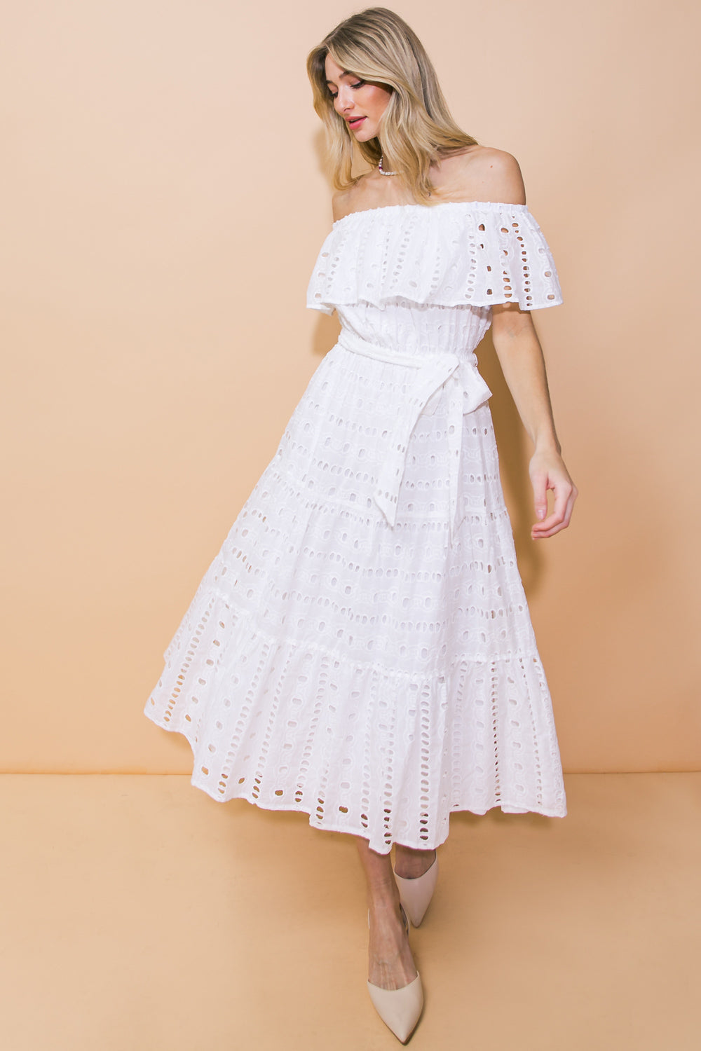 A woman in an off-the-shoulder white dress with eyelet detailing stands against a peach backdrop, gracefully displaying the knee-length garment while wearing beige heels and a pearl necklace.