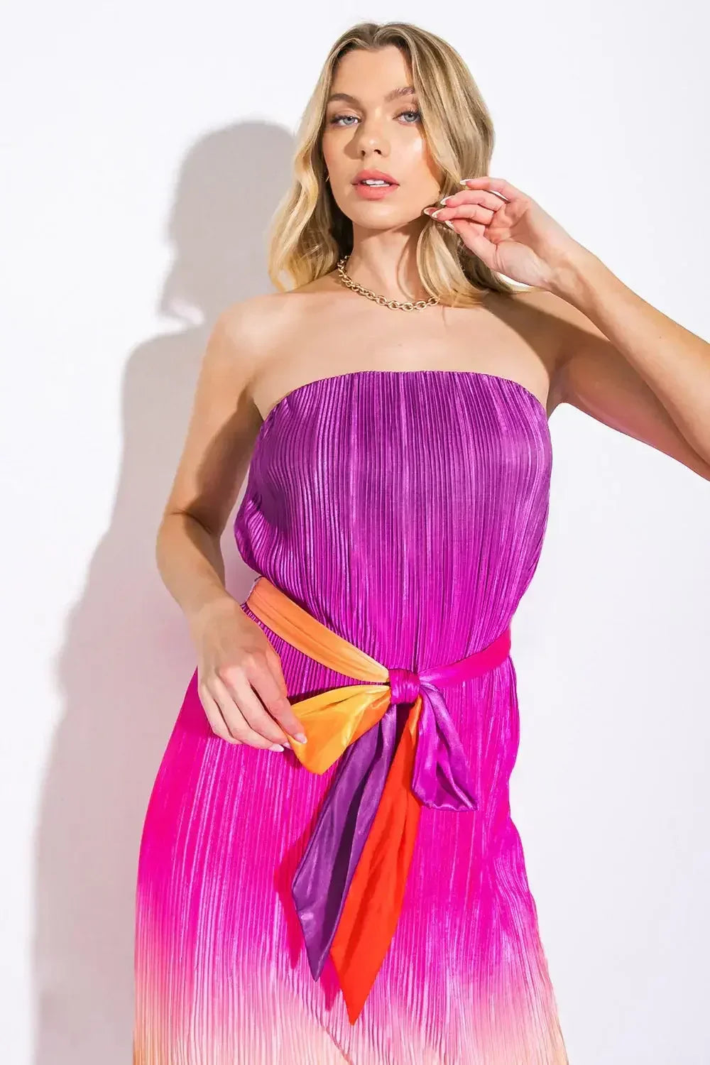 A woman poses in a strapless, pleated dress that transitions from purple to pink, tied with colorful ribbons. She is in a well-lit, neutral background, looking confidently at the camera.