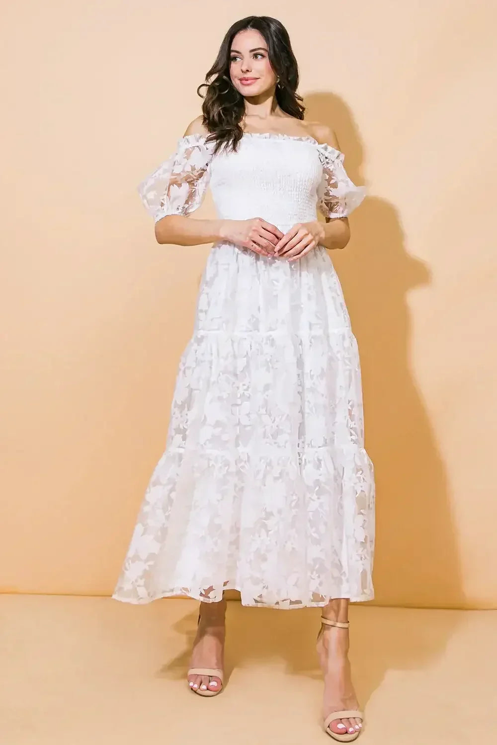 A woman in a white, off-the-shoulder dress with floral patterns stands elegantly against a soft orange background, holding her hands together and looking thoughtfully to her side.