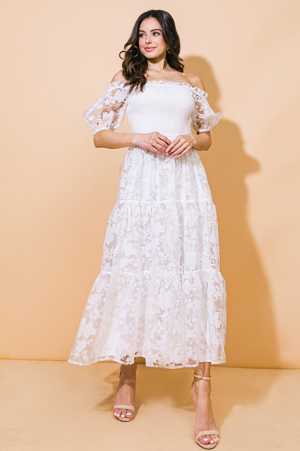 A woman in a white, off-the-shoulder dress with floral patterns stands elegantly against a soft orange background, holding her hands together and looking thoughtfully to her side.