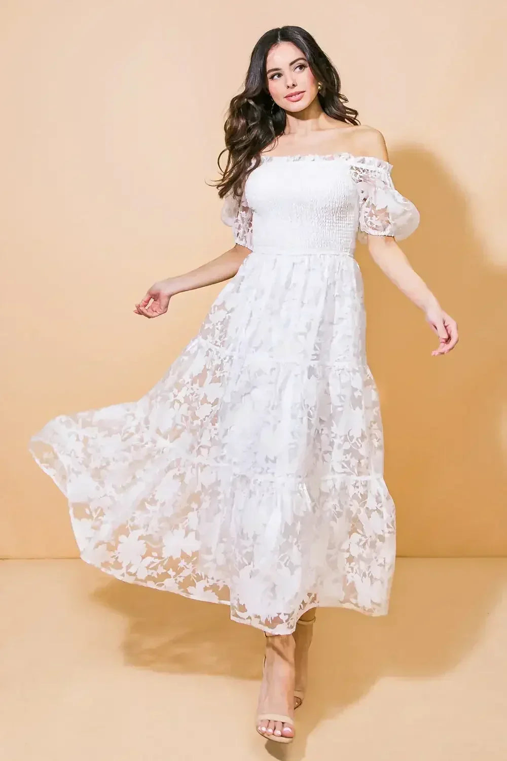 A woman in a flowing, off-shoulder white dress spins gracefully, revealing a sheer floral pattern. She stands against a soft, beige background, exuding a light, elegant demeanor.