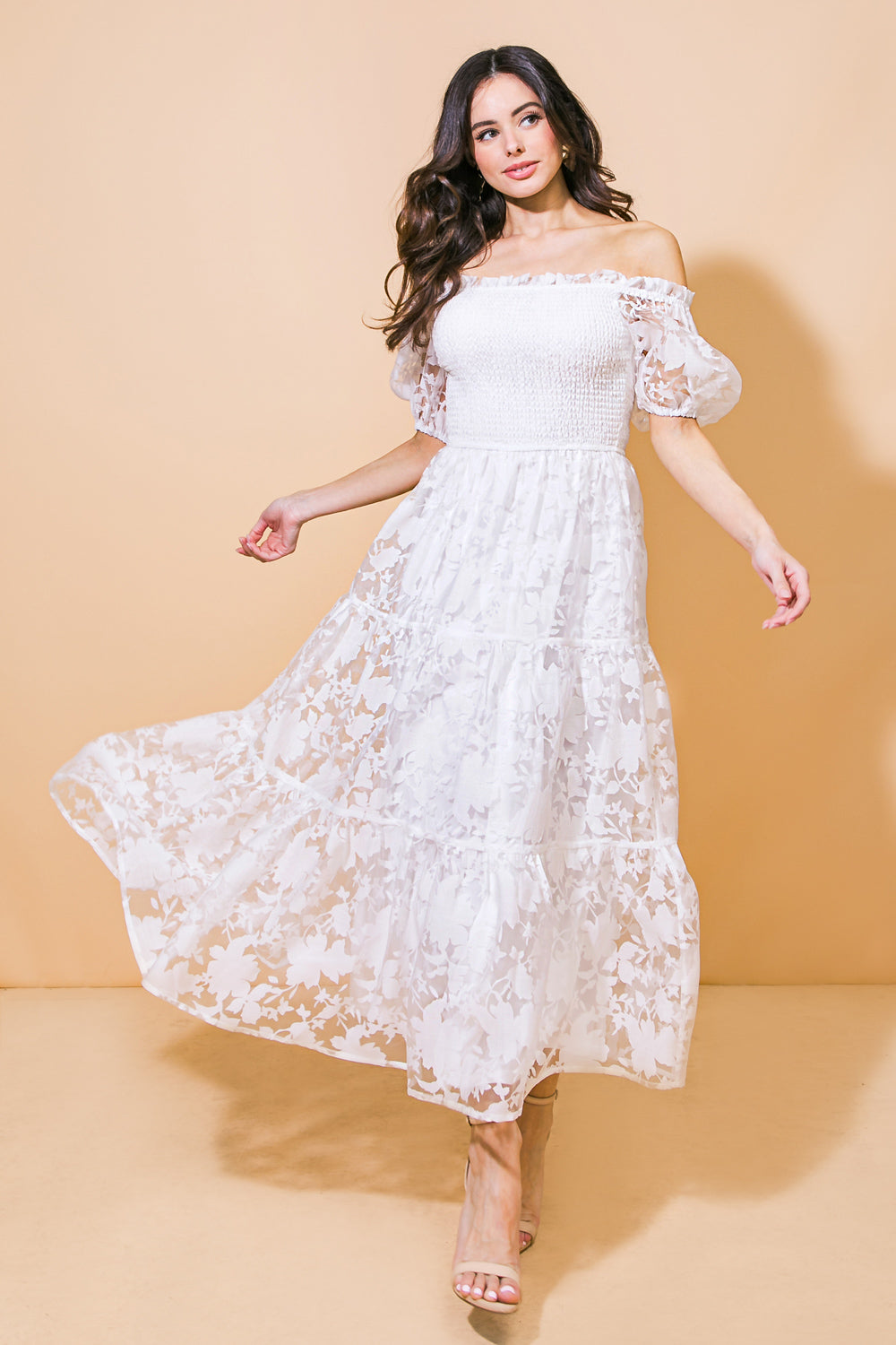 A woman in a flowing, off-shoulder white dress spins gracefully, revealing a sheer floral pattern. She stands against a soft, beige background, exuding a light, elegant demeanor.