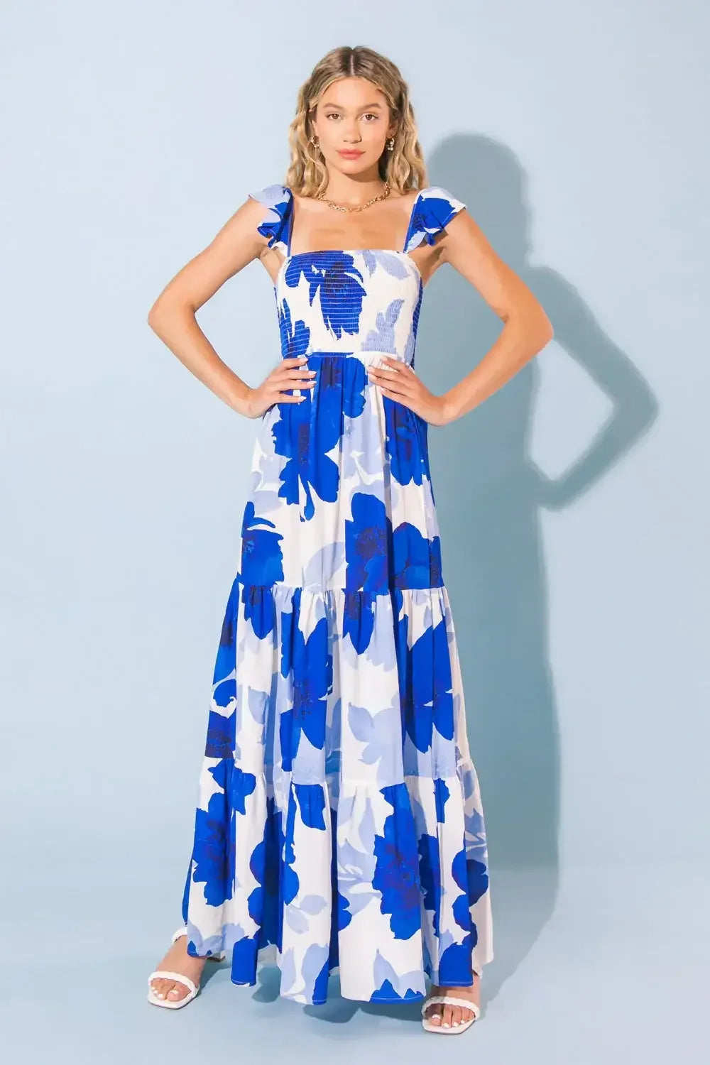 A woman stands confidently in a flowing, blue and white floral maxi dress with ruffled straps. The background is a simple light blue, enhancing the dress's vibrant colors.