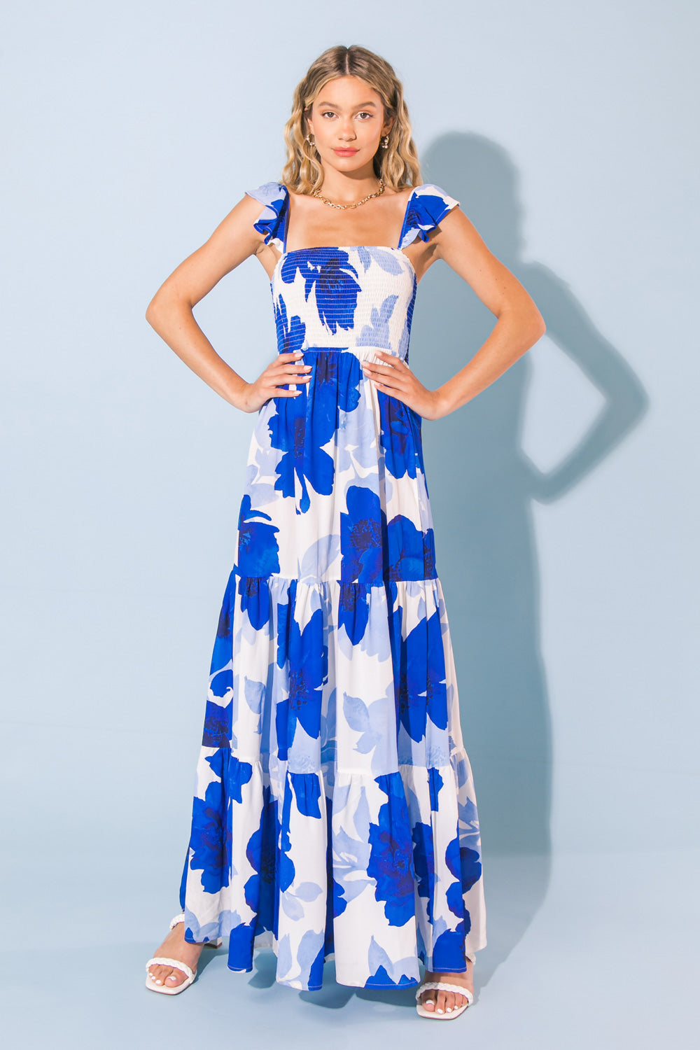 A woman stands confidently in a flowing, blue and white floral maxi dress with ruffled straps. The background is a simple light blue, enhancing the dress's vibrant colors.