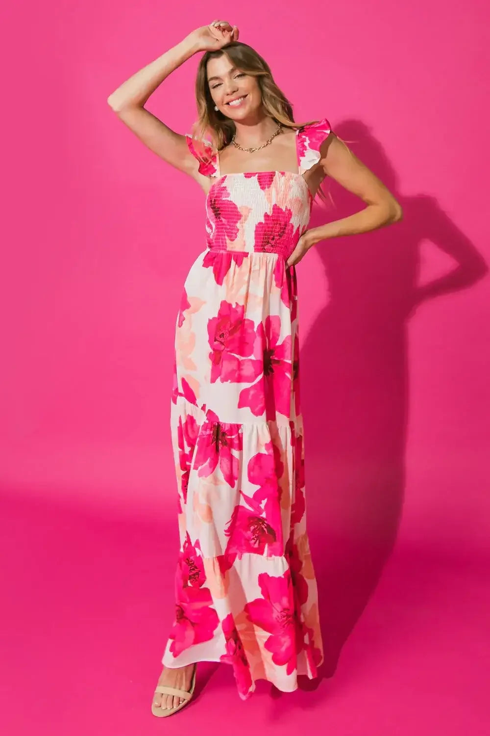 A woman wearing a long, floral-patterned dress poses playfully. She raises one arm and smiles, set against a vibrant pink background that enhances the dress's colors.