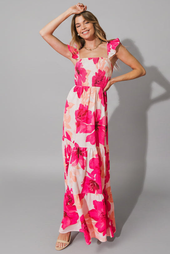 A woman wearing a long, floral-patterned dress poses playfully. She raises one arm and smiles, set against a vibrant pink background that enhances the dress's colors.