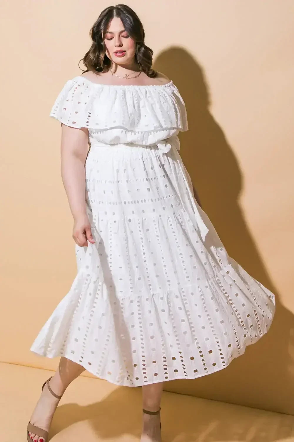 A person wearing a white off-shoulder dress with eyelet patterns stands gracefully, holding part of the dress, against a soft yellow backdrop, creating a warm and inviting atmosphere.