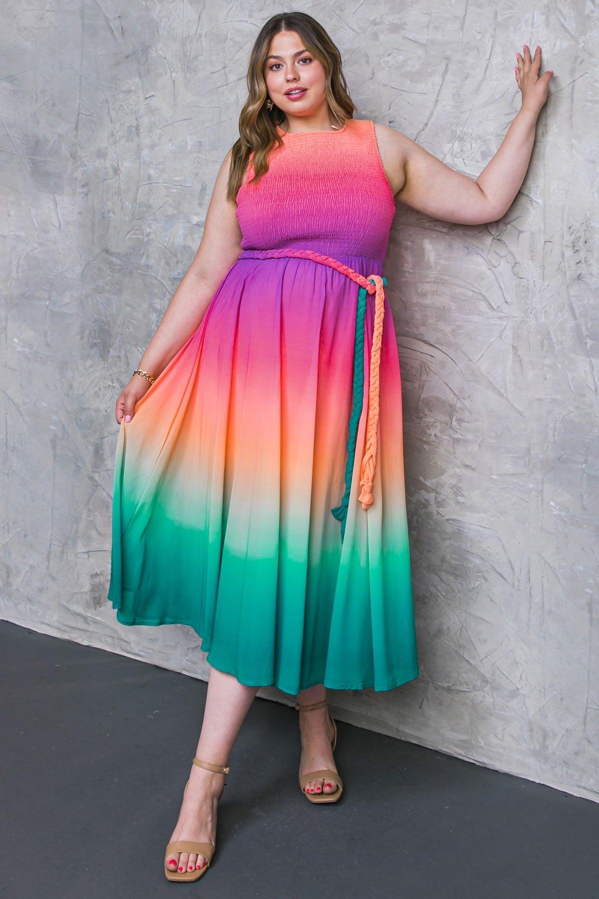 A woman in a colorful gradient dress stands against a textured gray wall, holding the fabric of her dress while wearing strappy sandals, showcasing a stylish look.