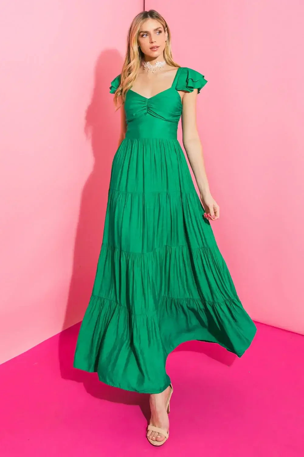 A model wearing a flowing green maxi dress with ruffled sleeves is standing against a pink backdrop. She appears poised, showcasing the dress’s tiers and fabric drape.