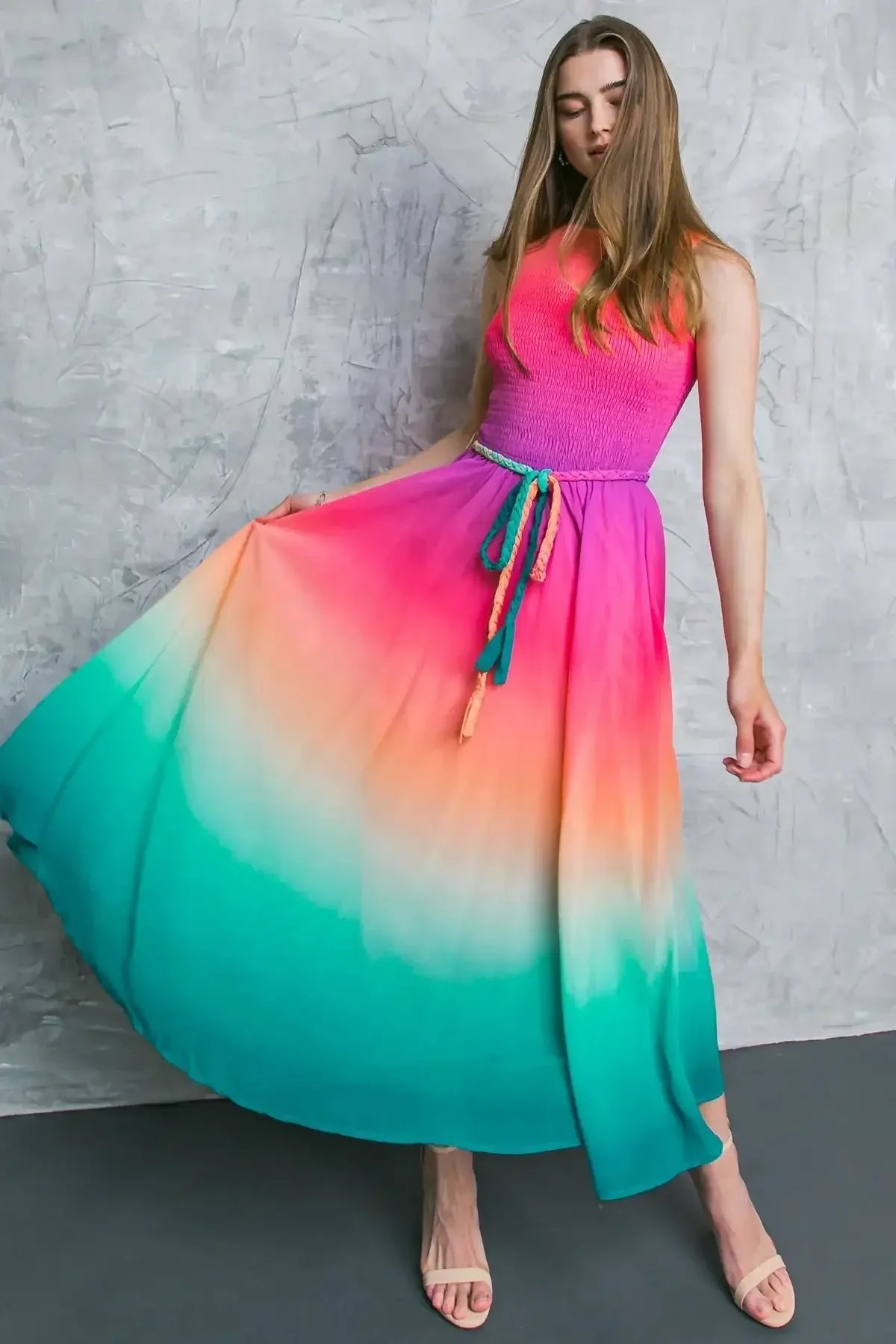 A woman wears a colorful, flowing dress with a gradient from pink to turquoise, elegantly lifting the skirt. She poses against a textured gray wall in a stylish setting.