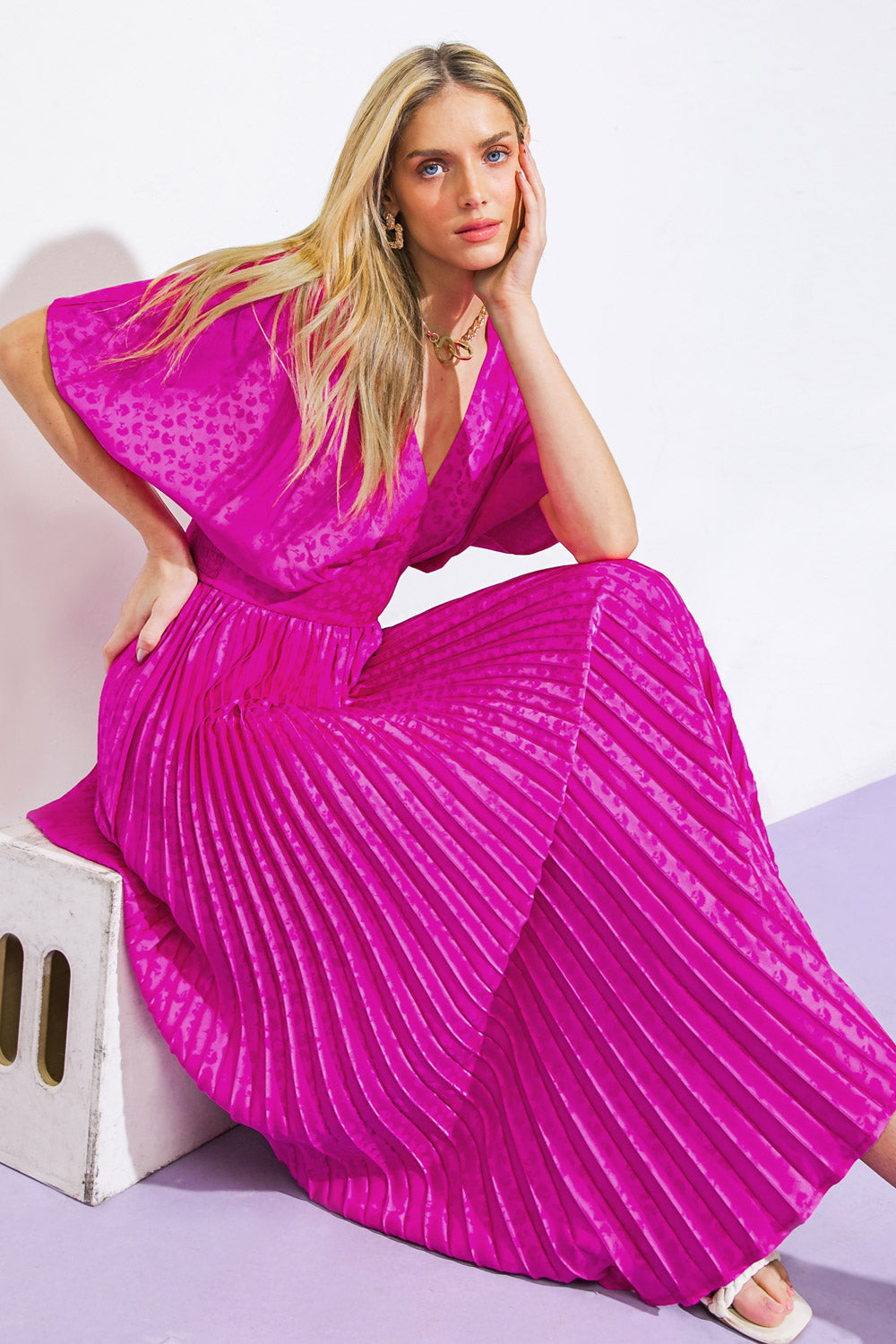 A woman sits elegantly on a light-colored stool, wearing a vibrant pink, pleated maxi dress. She rests her chin on her hand, posing against a minimalistic background.