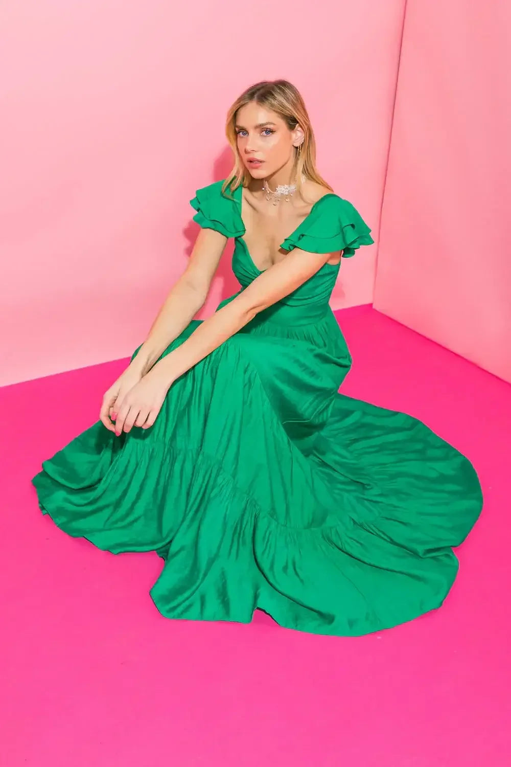 A woman sits gracefully in a flowing green dress on a vibrant pink floor. She poses with a serene expression, her hair styled softly, enhancing the colorful backdrop.