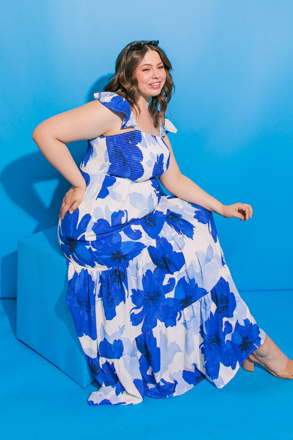 A woman sits on a blue platform, wearing a floral blue and white dress. She poses playfully with one hand on her hip and smiles, against a bright blue background.