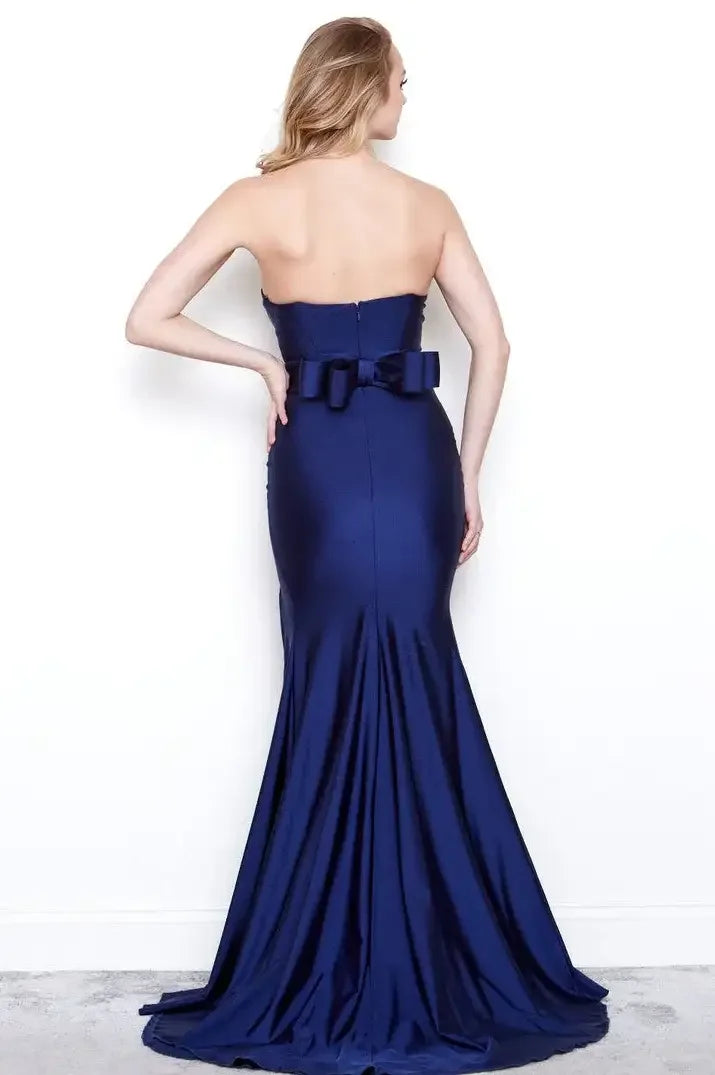 A woman is standing with her back to the viewer, wearing a fitted navy evening gown with a mermaid silhouette and a bow at the waist, against a plain white background.