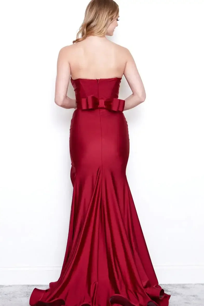A woman is wearing a floor-length, strapless red gown with a fitted bodice and a large bow at the back, standing against a plain white wall, showcasing the elegant draping of the material.