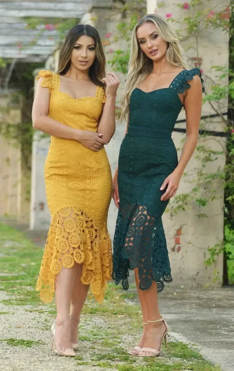 Two women stand side by side, dressed in elegant crochet dresses—one in yellow, the other in green. They pose outdoors on a pathway, surrounded by lush greenery and aged structures.