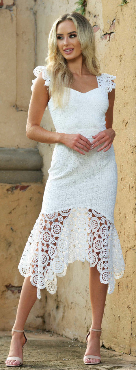 A woman in a white lace dress stands gracefully outdoors, smiling gently. The dress features intricate lace patterns and a high-low hem, against a rustic, textured wall.