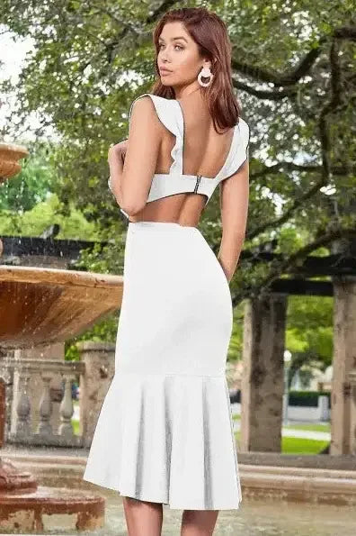 A woman stands with her back to the viewer, wearing a fitted white dress with a ruffled hem. She is in a garden-like setting near a fountain, surrounded by greenery.