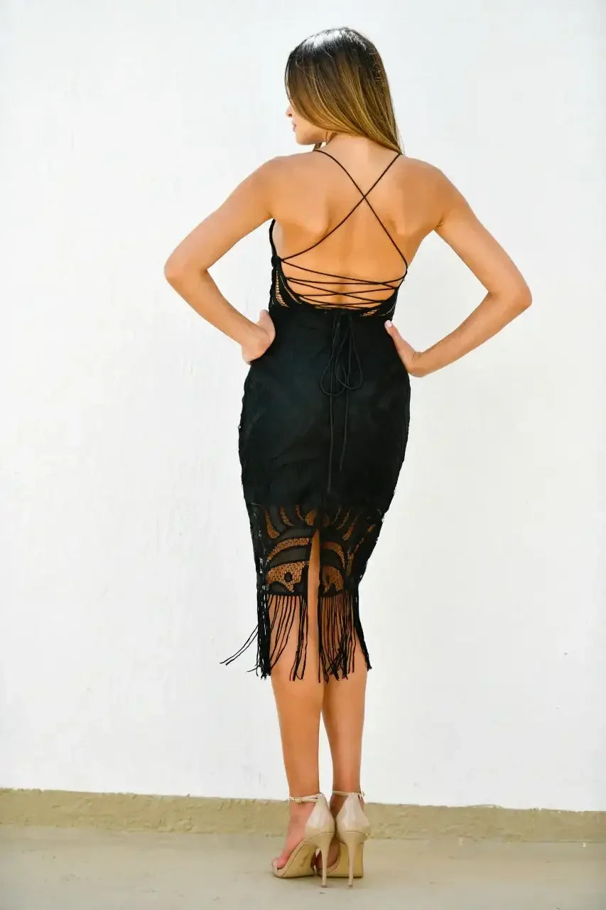 A woman stands with her back to the viewer, wearing a fitted black dress with a lace design and fringe. She poses against a plain white wall, showcasing her stylish footwear.