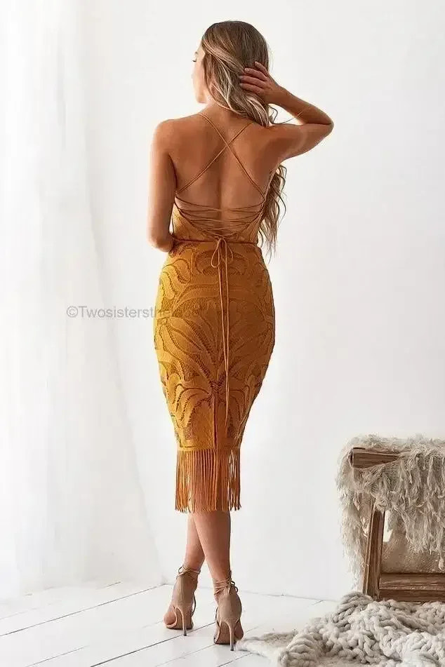 A woman stands with her back to the viewer, wearing a form-fitting, fringed mustard dress. Light streams in from large windows, illuminating a cozy, minimalist room with textured decor.